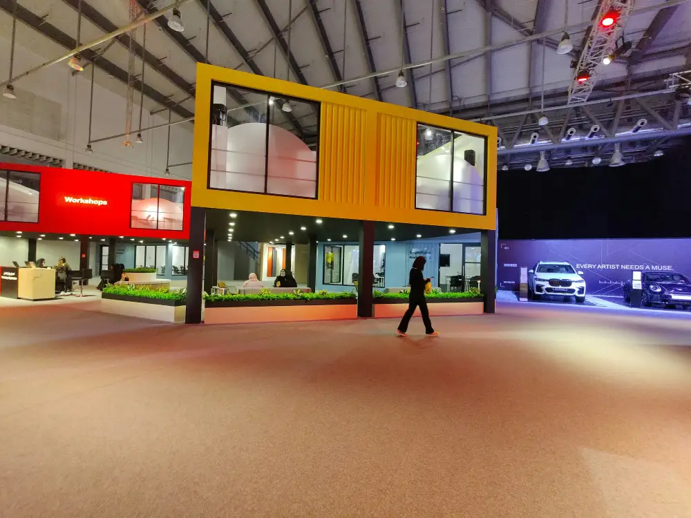 Visitors among illuminated exhibition stands beneath the trusses of a large indoor hall.