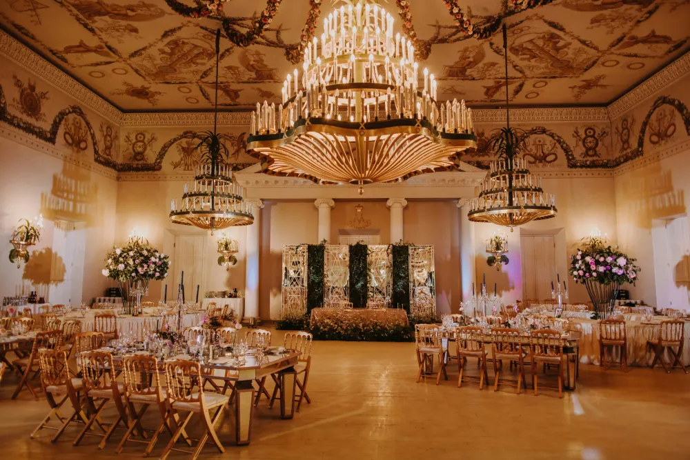 A reception hall with chandeliers, floral arrangements and gold chairs.